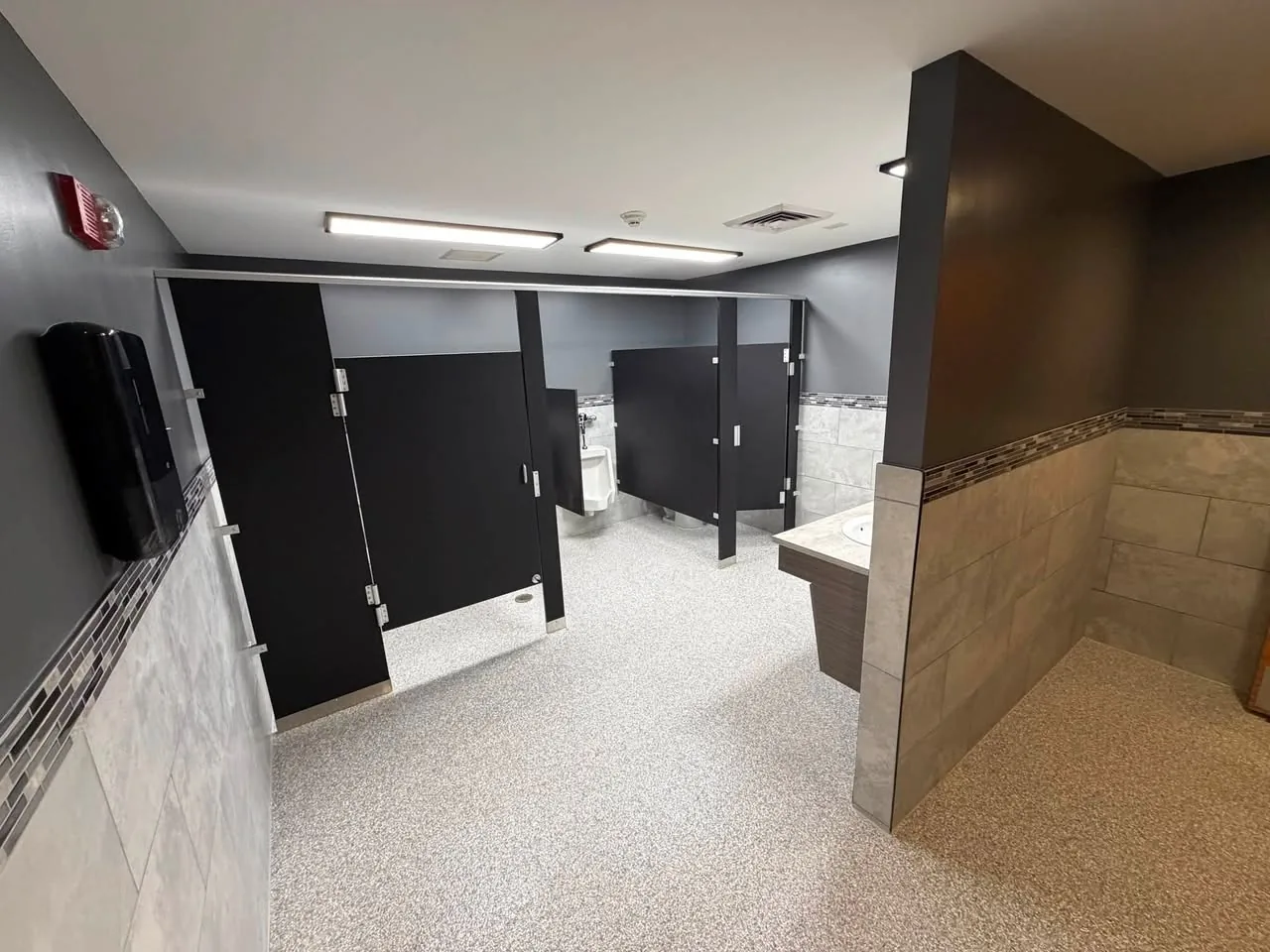 Remodeled banquet hall restroom with black stall partitions, gray wall tile, and a speckled gray floor