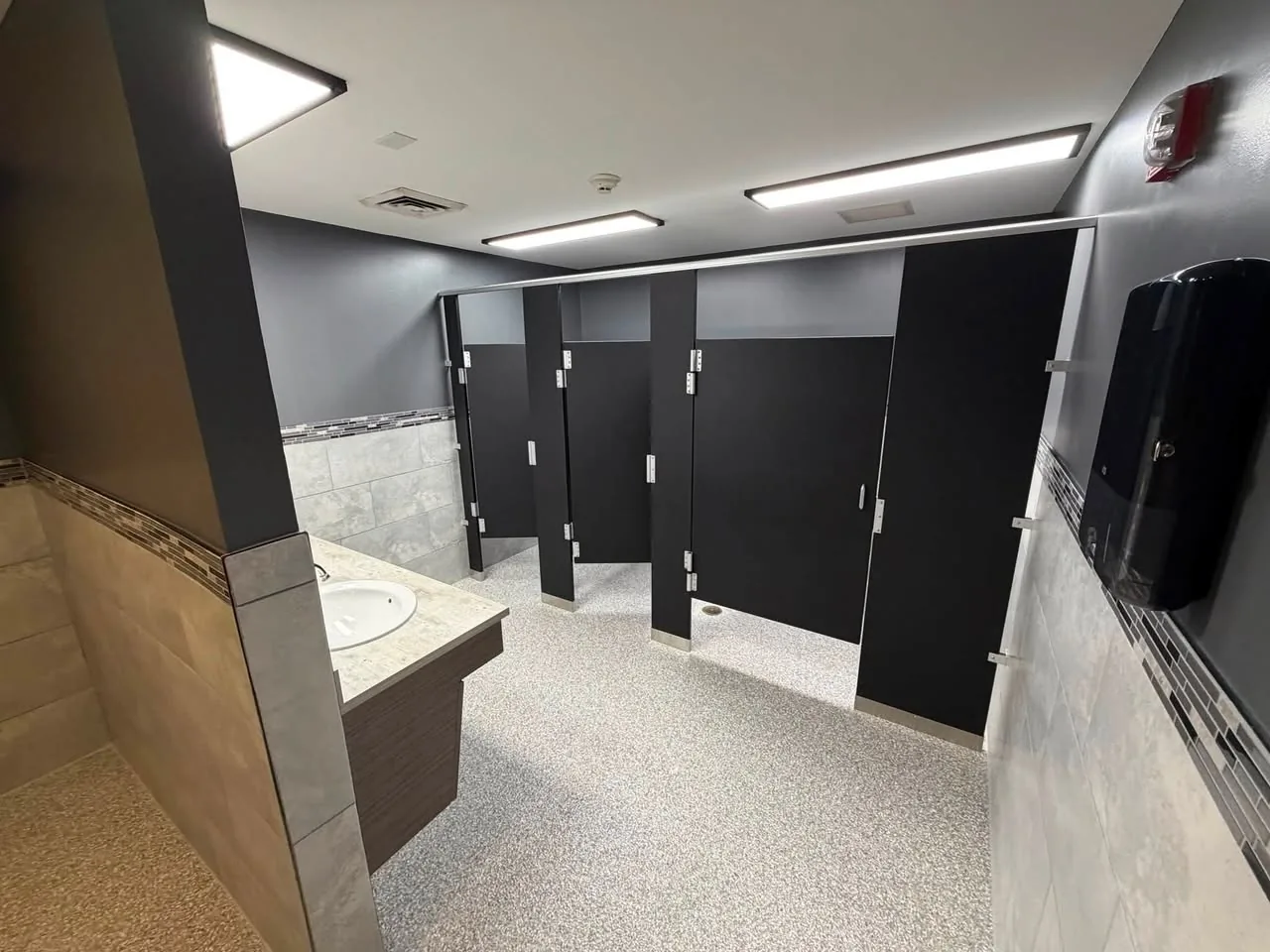 Remodeled restroom with black stall partitions, a mosaic tile band, and new lighting
